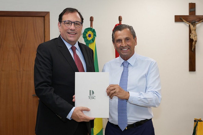 A imagem mostra duas pessoas, ambas homens, posando para uma foto em um ambiente formal. O homem à esquerda está vestindo um terno escuro com gravata vermelha e óculos, enquanto o homem à direita está usando uma camisa social azul clara com gravata azul. Eles estão segurando juntos um documento que tem o logotipo e as letras "TJSC" impressas na capa. Ao fundo, há uma bandeira do Brasil e outra bandeira, além de um crucifixo pendurado na parede. O ambiente parece ser um escritório ou sala de reuniões.