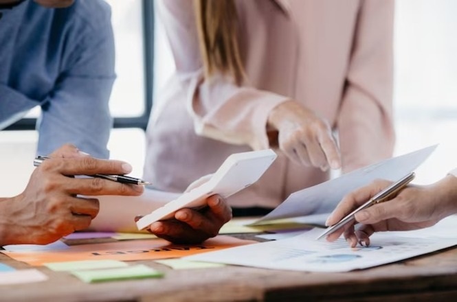A imagem mostra três pessoas em uma reunião ou discussão de trabalho. Elas estão em volta de uma mesa, onde há papéis e notas adesivas coloridas espalhadas. Uma das pessoas está segurando uma calculadora, enquanto as outras duas estão apontando para os papéis com canetas. A cena sugere que estão analisando ou discutindo informações, possivelmente relacionadas a números ou dados. As roupas das pessoas são casuais, e a imagem transmite uma atmosfera de colaboração e foco.