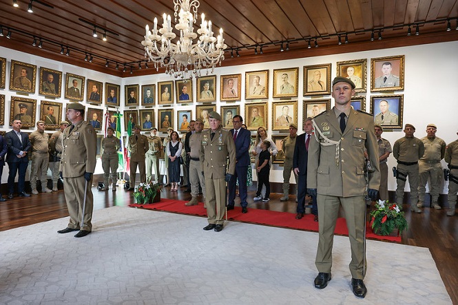 A imagem mostra uma cerimônia militar em uma sala com paredes cobertas por retratos de pessoas emolduradas. No centro da sala, há três militares em uniforme formal, com um deles em destaque na frente. Eles estão em pé sobre um tapete claro, e há um lustre pendurado no teto de madeira. Ao fundo, há um grupo de pessoas, incluindo outros militares e civis, alguns dos quais estão em uniforme militar e outros em trajes formais. Há também bandeiras e arranjos de flores no ambiente. A atmosfera é formal e solene.