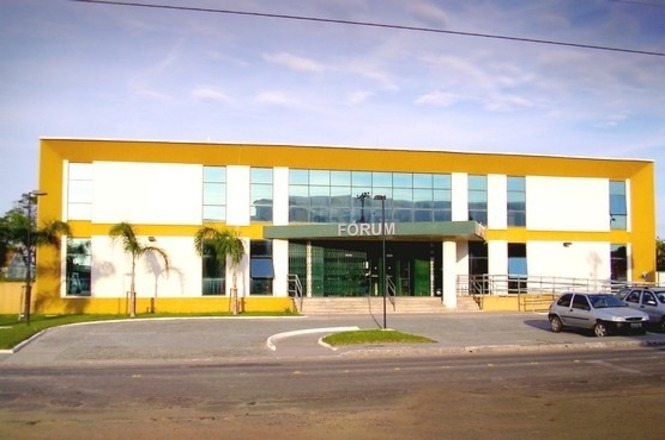 A imagem mostra a fachada de um prédio moderno com dois andares, identificado como "FÓRUM" em letras grandes acima da entrada principal. A construção é predominantemente branca com detalhes em amarelo nas bordas. Há várias janelas de vidro azuladas que refletem o céu. Na frente do prédio, há uma área pavimentada com algumas palmeiras e um estacionamento onde estão estacionados dois carros. O céu está parcialmente nublado.