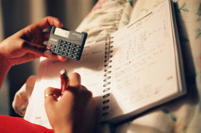 A imagem mostra uma pessoa segurando uma calculadora na mão esquerda enquanto escreve em um caderno espiral com a mão direita. A calculadora é pequena e parece ser do tipo portátil. O caderno está aberto em uma página que contém várias anotações e cálculos escritos à mão. A pessoa está usando uma caneta vermelha para escrever. O ambiente parece ser confortável, possivelmente um quarto, já que há um cobertor ou edredom floral visível ao fundo. A iluminação é suave, sugerindo um ambiente tranquilo e relaxante.