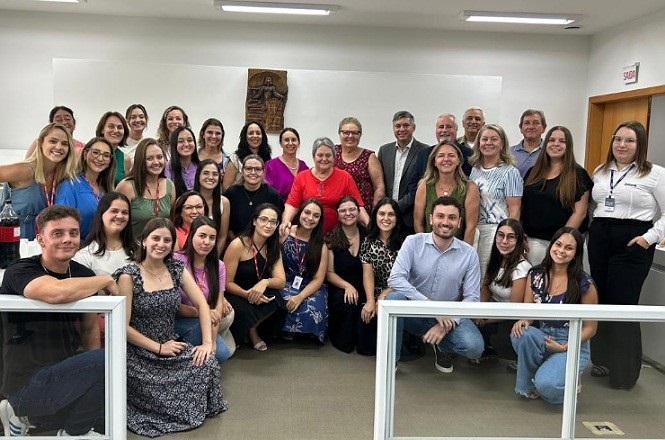 A imagem mostra um grupo de aproximadamente 36 pessoas posando para uma foto em uma sala. A maioria das pessoas está sorrindo e parece estar em um ambiente de trabalho ou reunião. Há uma mistura de homens e mulheres, e a maioria está vestida de maneira casual ou semi-formal. No fundo, há uma parede branca com uma peça de arte pendurada. A sala tem iluminação fluorescente e há uma porta à direita com uma placa de "saída" acima. Algumas pessoas estão sentadas na frente, enquanto outras estão em pé atrás.
