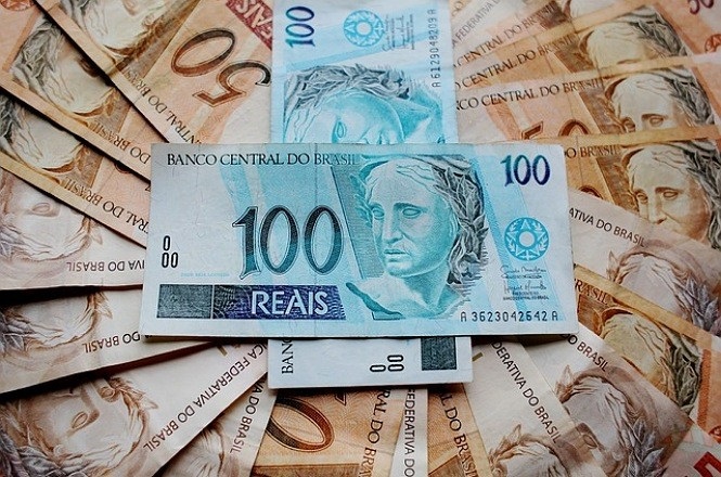 A imagem mostra várias notas de dinheiro brasileiro espalhadas. No centro, há uma nota de 100 reais, que é azul e tem a imagem de uma escultura representando a República. Ao fundo, há várias notas de 50 reais, que são de cor marrom e também têm a imagem da escultura da República. As notas estão dispostas de forma sobreposta, criando um efeito de leque.