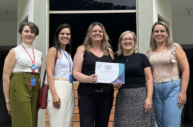 A imagem mostra cinco mulheres em pé, lado a lado, sorrindo para a câmera. A mulher no centro está segurando um certificado. Elas estão em frente a um prédio com uma porta de vidro ao fundo. As mulheres estão vestidas de maneira casual e parecem estar em um ambiente externo, possivelmente em um evento ou cerimônia de entrega de certificados. O certificado que a mulher no centro segura tem o título "Certificado Escola CIDADÃ".