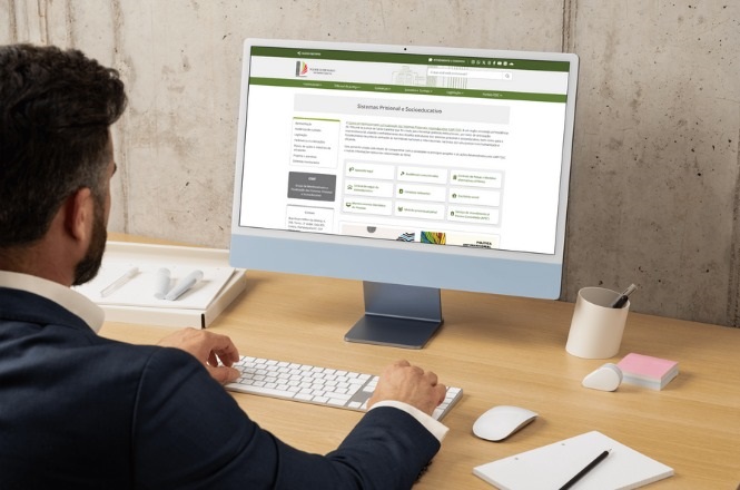 A imagem mostra um homem sentado em frente a um computador em uma mesa de madeira. Ele está usando um terno escuro e está olhando para a tela do computador. Na tela, há uma página da web aberta que parece ser um sistema ou portal online, com um cabeçalho verde e várias seções e botões organizados em caixas. Há um teclado e um mouse branco sobre a mesa, além de um bloco de notas com uma caneta e uma caneca branca. A parede ao fundo é de concreto aparente.