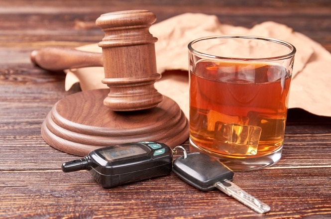 A imagem mostra um martelo de juiz de madeira ao lado de um copo de bebida alcoólica, que parece ser uísque, sobre uma superfície de madeira. Ao lado do copo, há um chaveiro com uma chave de carro e um dispositivo que parece ser um bafômetro portátil. A composição sugere uma mensagem sobre a relação entre dirigir sob a influência de álcool e as consequências legais.