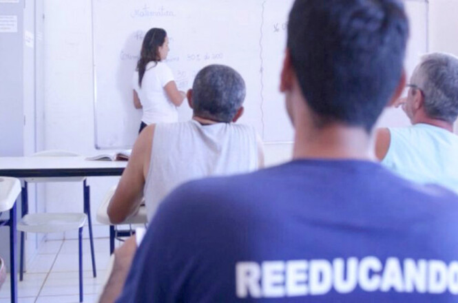 A imagem mostra uma sala de aula com algumas pessoas sentadas em carteiras, voltadas para um quadro branco. No quadro, há uma mulher escrevendo algo. Ela está de pé, de costas para a câmera. No primeiro plano, há um homem usando uma camiseta azul com a palavra "REEDUCANDO" escrita nas costas. A sala parece ser simples, com paredes brancas e cadeiras de plástico. A atmosfera sugere um ambiente de ensino, possivelmente em um contexto de reeducação ou reintegração social.