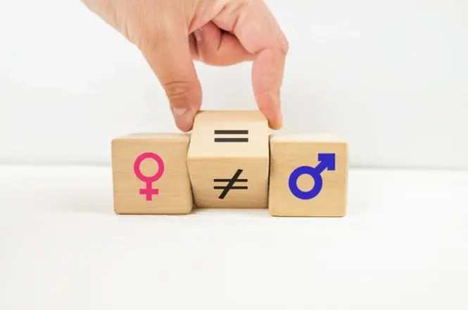 A imagem mostra três blocos de madeira lado a lado. No bloco à esquerda, há o símbolo feminino em rosa. No bloco à direita, há o símbolo masculino em azul. O bloco do meio tem dois símbolos: o de igualdade (=) na parte superior e o de desigualdade (≠) na parte inferior. Uma mão está segurando o bloco do meio, sugerindo a escolha entre igualdade e desigualdade entre os gêneros.