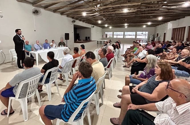 Uma reunião em um salão amplo com várias pessoas sentadas em cadeiras de plástico branco. À esquerda, um homem de terno preto está em pé, falando ao microfone. À frente dele, há uma mesa com várias pessoas sentadas, possivelmente uma mesa de palestrantes. O salão tem um teto de madeira exposta e iluminação de lâmpadas embutidas. As pessoas na audiência estão atentas, algumas com os braços cruzados, outras com as mãos no queixo. Ao fundo, há janelas grandes que permitem a entrada de luz natural.