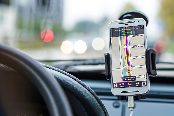 Um carro com um smartphone preso em um suporte no painel. O smartphone está exibindo um aplicativo de navegação GPS, mostrando um mapa com uma rota destacada em roxo. No canto inferior da tela do telefone, há informações de navegação, como a hora atual (10:32), a distância restante (1,9 km) e a velocidade (43 km/h). O volante do carro é visível à esquerda, e a estrada à frente está desfocada, com algumas luzes de veículos ao fundo.