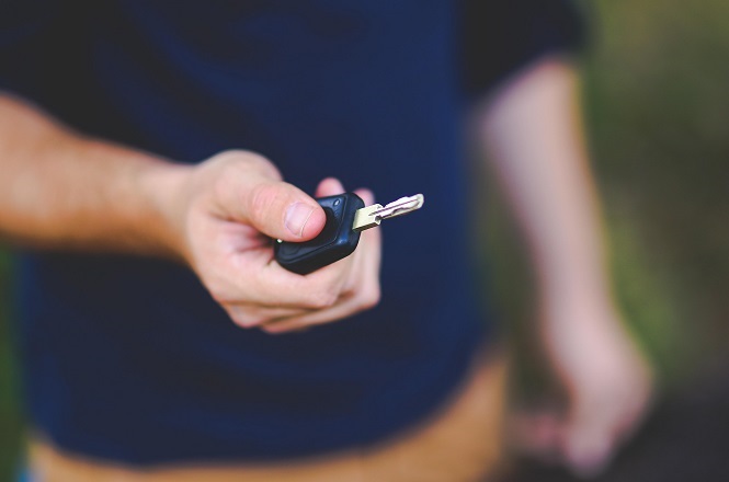 Uma mão segurando uma chave de carro com um controle remoto preto. A pessoa está vestindo uma camisa azul escura e calças marrons. O fundo está desfocado, destacando a chave e a mão.