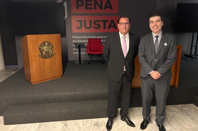 Dois homens de terno estão posando para uma foto em um ambiente formal. O homem à esquerda usa óculos, um terno preto, camisa branca e gravata rosa clara. O homem à direita veste um terno cinza escuro, camisa azul clara e gravata cinza. Ambos estão sorrindo. Atrás deles, há um palco com um púlpito de madeira com o brasão do Brasil. No fundo, há uma parede com as palavras "PENA JUSTA" em destaque. Há também uma cadeira vermelha e um fundo escuro com texto em letras menores, que não é totalmente legível.