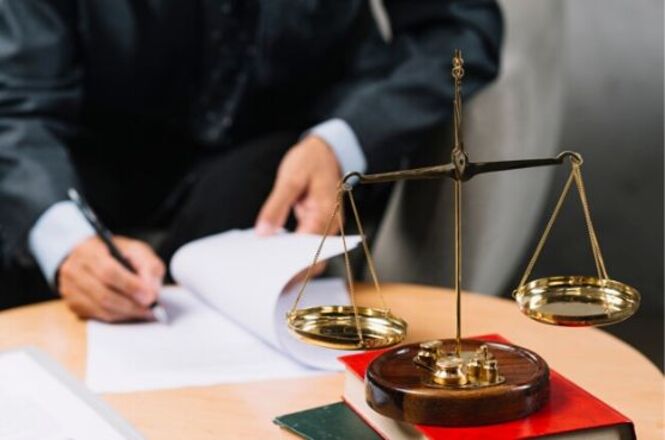 Uma balança de justiça dourada está em destaque sobre uma mesa. Ao lado, há um livro vermelho e outro verde empilhados. Ao fundo, uma pessoa vestindo um terno preto está assinando um documento. A cena sugere um ambiente jurídico ou de escritório de advocacia.