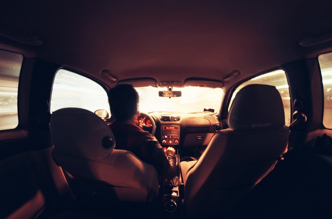 A imagem mostra o interior de um carro em movimento à noite, vista do banco traseiro. O motorista, que está de costas para a câmera, segura o volante enquanto olha para a estrada. O painel do carro está iluminado com luzes avermelhadas, e há um efeito de movimento nas janelas, onde as luzes externas aparecem borradas, criando uma sensação de velocidade. O interior do carro tem tons quentes, com os bancos dianteiros cobertos de um tecido claro. O para-brisa mostra um brilho intenso à frente, possivelmente de faróis ou luzes urbanas.