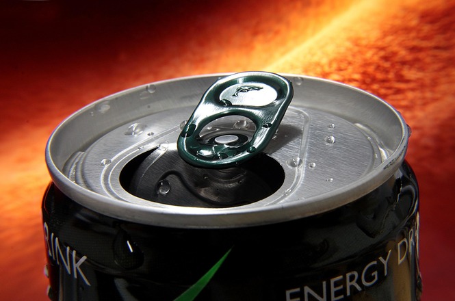A imagem mostra uma lata de energético aberta, com gotas de líquido espalhadas sobre sua superfície metálica. A lingueta da lata é de cor verde e está levantada, revelando a abertura por onde a bebida pode ser consumida. O fundo da imagem tem um tom quente e vibrante, com um efeito que lembra um líquido ou gás em movimento, sugerindo energia e dinamismo. A lata em si é preta, com detalhes em verde e branco, transmitindo uma aparência moderna e intensa.