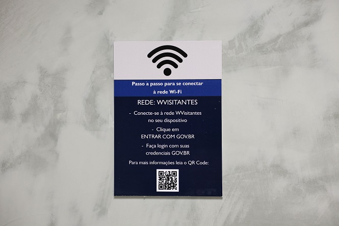 A imagem mostra um cartaz com instruções para se conectar a uma rede Wi-Fi. Na parte superior, há um ícone de sinal Wi-Fi. Abaixo, as instruções são divididas em etapas: conectar-se à rede "WVvisitantes" no dispositivo, clicar em "ENTRAR COM GOV.BR" e fazer login com as credenciais GOV.BR. Há também um QR Code no final para mais informações. O fundo do cartaz é dividido em cores branca e azul.