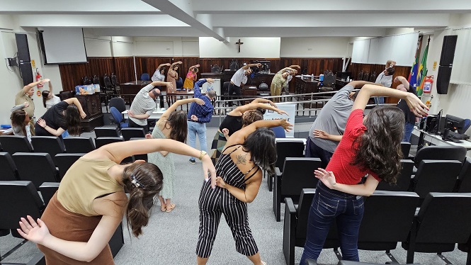 A imagem mostra um grupo de pessoas em uma sala, provavelmente um auditório ou sala de aula, realizando exercícios de alongamento. Elas estão inclinadas lateralmente, com um braço levantado acima da cabeça. O ambiente tem cadeiras dispostas em fileiras, e um crucifixo pode ser visto na parede ao fundo, indicando que o local pode ser uma instituição educacional ou religiosa. Há também bandeiras visíveis no canto direito da imagem.