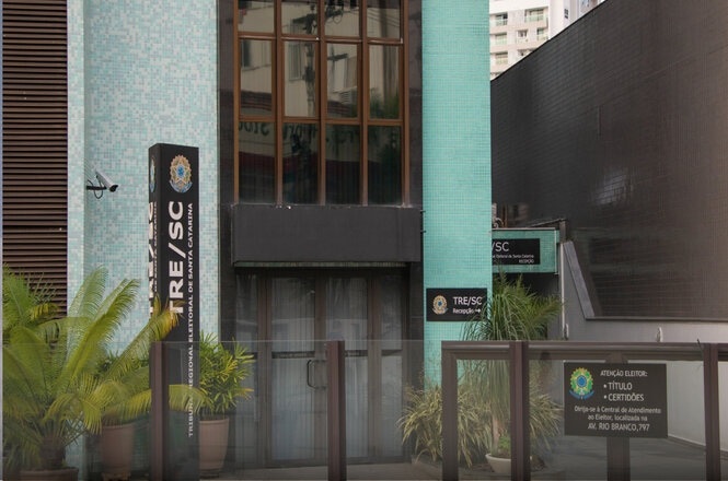 A imagem mostra a fachada de um edifício com azulejos azuis claros, que pertence ao TRE/SC, o Tribunal Regional Eleitoral de Santa Catarina. Há placas indicando que é o setor de recepção e atenção ao público para títulos e certidões. Na frente, há uma grade e algumas plantas em vasos. O design do prédio é moderno e urbano, e parece estar localizado em uma área movimentada com prédios ao fundo.