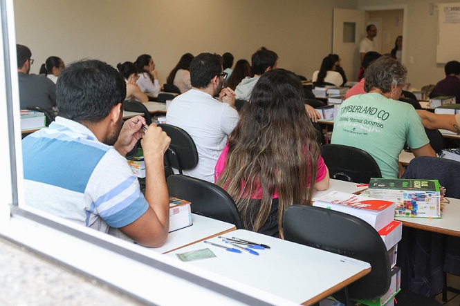 A imagem mostra uma sala de aula com várias pessoas sentadas em carteiras, assistindo a uma apresentação ou aula. Eles parecem estar prestando atenção ao que está sendo apresentado na frente da sala. Há livros e materiais sobre as mesas, e a maioria das pessoas está sentada de costas para a câmera. A imagem é tirada de fora da sala, através de uma janela.