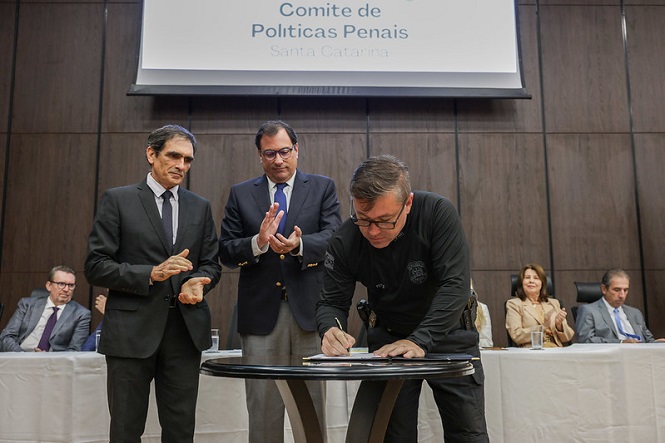 A imagem mostra uma cerimônia oficial com várias pessoas. No centro, há um homem assinando um documento sobre uma mesa circular. Atrás dele, duas outras pessoas estão em pé e aplaudindo, enquanto outras estão sentadas em uma mesa ao fundo. Acima, há uma faixa ou quadro com os dizeres "Comitê de Políticas Penais Santa Catarina". O ambiente parece ser uma sala de conferências ou um auditório.
