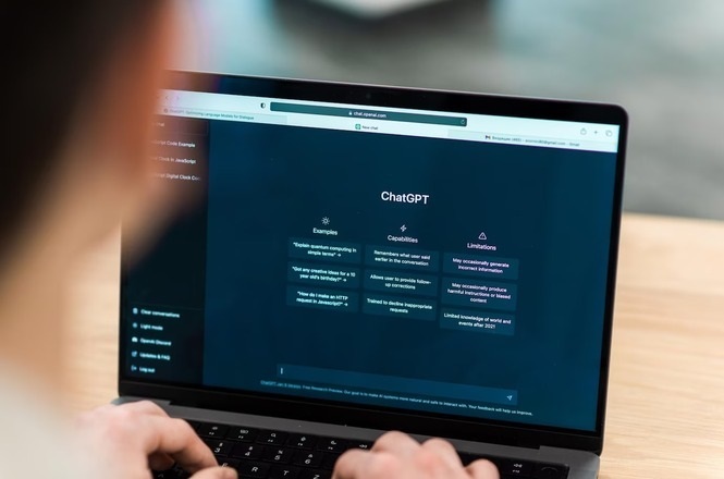 A imagem mostra uma pessoa usando um laptop que exibe a interface do ChatGPT na tela. A tela do computador tem um fundo escuro com texto em branco, dividida em três seções principais: "Exemplos", "Capacidades" e "Limitações". As mãos da pessoa estão no teclado, sugerindo que está interagindo com o computador. A cena está em um ambiente de escritório ou similar, com uma superfície de madeira abaixo do laptop.
