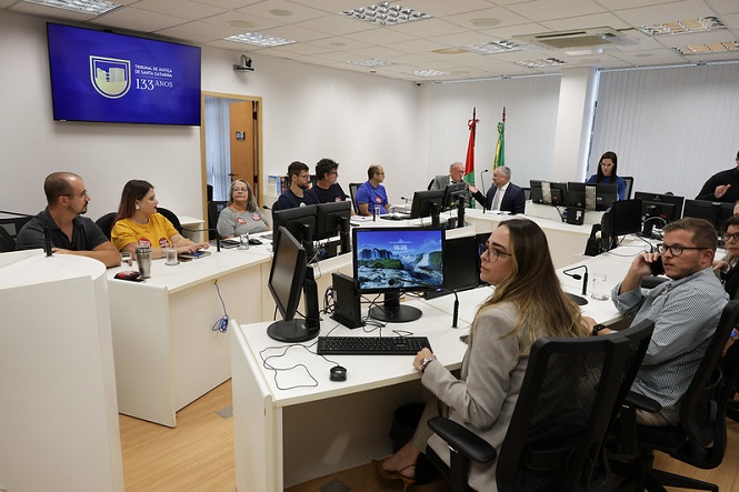 A imagem mostra uma sala de conferência ou reunião, onde várias pessoas estão sentadas em torno de mesas dispostas em forma de U. Elas estão usando computadores e prestando atenção ao que parece ser uma apresentação ou discussão. Na parede, há um grande monitor exibindo um logotipo e "133 anos". Há bandeiras em um dos cantos da sala, sugerindo um ambiente institucional ou governamental. A sala é bem iluminada e tem um ambiente moderno e profissional.