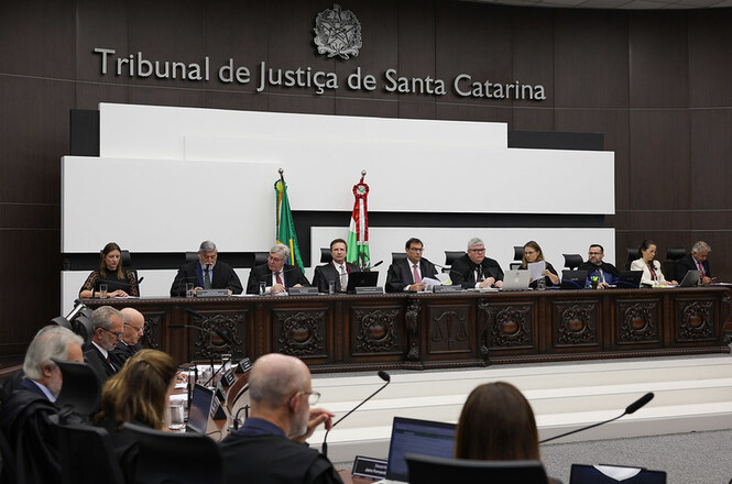 A imagem mostra uma sessão no Tribunal de Justiça de Santa Catarina. Há várias pessoas sentadas atrás de uma mesa comprida, possivelmente juízes ou membros do tribunal, com bandeiras ao fundo. O ambiente tem um aspecto formal e várias pessoas estão utilizando laptops e papéis, sugerindo um trabalho judicial em andamento. A decoração da mesa é elaborada, e há microfones à disposição dos participantes.