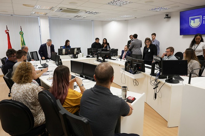A imagem mostra uma sala de reuniões com várias pessoas sentadas e em pé ao redor de uma mesa em formato de "U". Há computadores e monitores na mesa, e algumas pessoas estão interagindo ou trabalhando nelas. Na parede, há uma tela grande exibindo um logotipo. A sala tem um ambiente corporativo, com luminárias no teto e bandeiras em um canto. A atmosfera parece ser formal, sugerindo uma reunião ou atividade relacionada a negócios ou educacional.