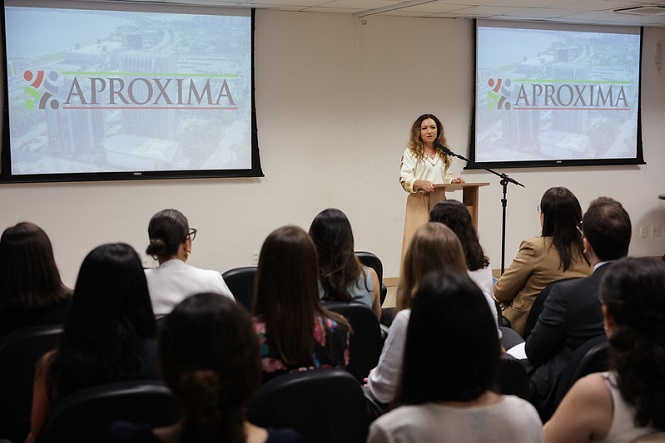 A imagem mostra uma mulher em pé, falando em um palco ou tribuna, diante de um público sentado. Atrás dela, há dois projetores com o logotipo "APROXIMA" exibido em uma tela. A audiência é composta por várias pessoas, e a sala parece ser um ambiente de conferência ou reunião. A mulher parece estar apresentando ou discursando sobre um tema relacionado ao que está projetado.