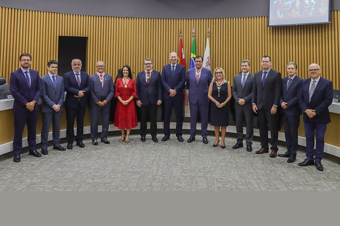 A imagem mostra um grupo de pessoas em pé, enfileiradas em um ambiente interno, possivelmente uma sala de reuniões ou conferência. Há bandeiras ao fundo e uma tela no canto superior. As pessoas estão vestidas de maneira formal, a maioria usando ternos e trajes sociais. Algumas delas usam medalhas no pescoço, indicando possível participação em um evento oficial ou cerimônia. O chão é coberto por um carpete cinza e as paredes têm um revestimento que parece de madeira.