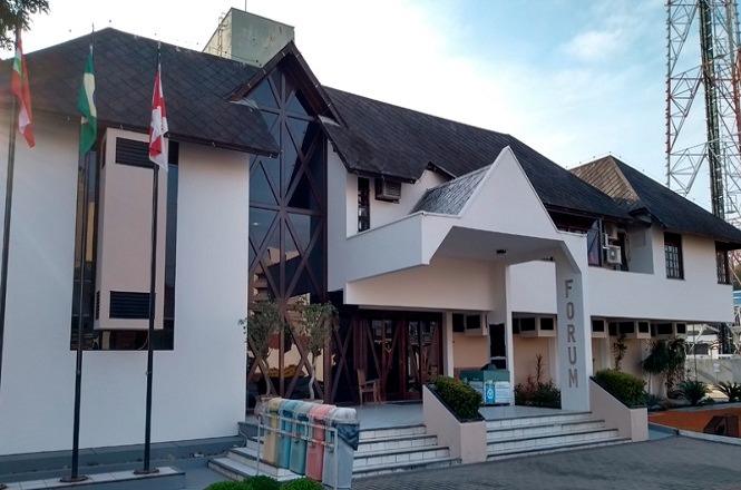 Prédio com arquitetura moderna, com telhado inclinado e janelas de vidro em formato triangular. Na entrada, há uma escadaria com corrimão e uma cobertura em forma de triângulo. Ao lado da escada, há um letreiro vertical com a palavra "FÓRUM". Na frente do prédio, há três mastros com bandeiras. O ambiente ao redor é limpo e bem cuidado, com algumas plantas decorativas.