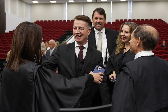 Um grupo de pessoas está em um ambiente formal, possivelmente um auditório ou tribunal, com cadeiras vermelhas ao fundo. As pessoas estão vestidas com trajes formais, como togas e ternos. No centro, um homem de terno escuro e gravata está sorrindo e cumprimentando uma mulher de costas, que também está usando uma toga. Ao redor deles, há outras pessoas sorrindo, incluindo uma mulher loira e um homem de barba. O ambiente parece ser iluminado e espaçoso.