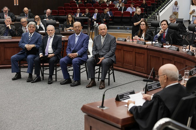 Sala de tribunal com várias pessoas presentes. Na frente, quatro homens estão sentados em cadeiras, todos vestidos formalmente com ternos. Atrás deles, há uma bancada em formato de "U" com várias pessoas sentadas, algumas usando togas, sugerindo que são juízes ou advogados. No centro da imagem, um homem careca, também de toga, está sentado em uma cadeira maior, possivelmente presidindo a sessão. Ao fundo, há mais pessoas sentadas em cadeiras vermelhas, observando a sessão. O ambiente é formal e parece ser uma audiência ou julgamento