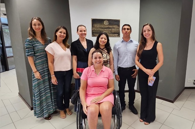Um grupo de sete pessoas está posando para uma foto em um ambiente interno. No centro, uma mulher está sentada em uma cadeira de rodas, vestindo um vestido rosa. Ao redor dela, há três mulheres à esquerda e duas mulheres e um homem à direita. As mulheres estão vestidas com roupas casuais e elegantes, enquanto o homem usa uma camisa azul clara e calças escuras. Ao fundo, há uma placa na parede com inscrições, mas o texto não é legível na imagem. O ambiente parece ser um local institucional ou de escritório, com paredes cinza e piso claro.