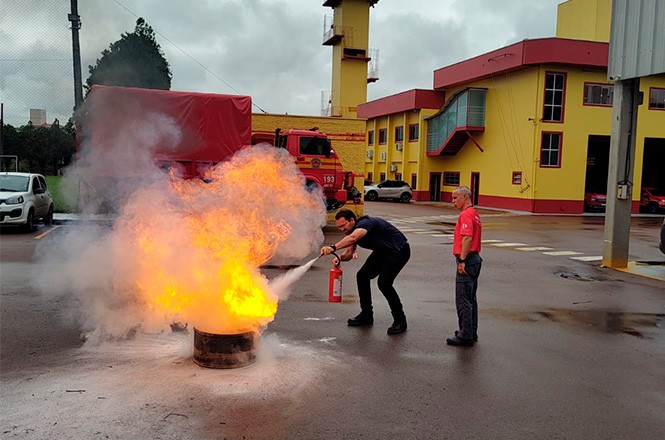 Um homem está usando um extintor de incêndio para apagar chamas que saem de um recipiente no chão. Ele está sendo supervisionado por outro homem, que está de pé ao lado dele. Ambos estão em um pátio de um corpo de bombeiros, com um caminhão de bombeiros vermelho estacionado ao fundo. O prédio do corpo de bombeiros é amarelo com detalhes em vermelho. O céu está nublado, indicando um dia possivelmente chuvoso