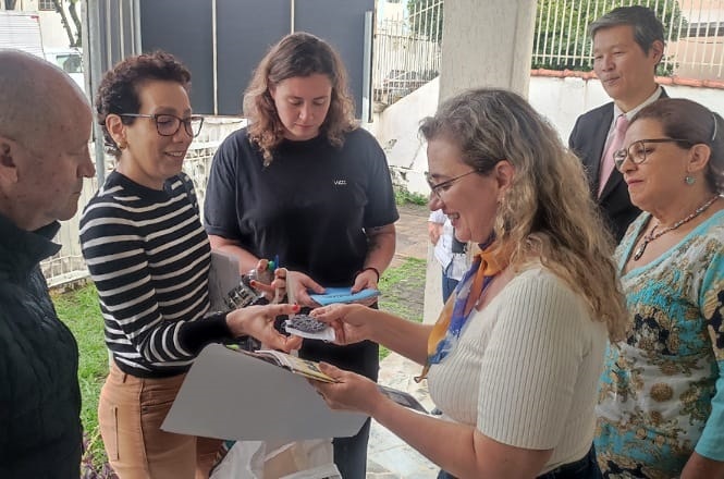 Um grupo de seis pessoas está reunido em um espaço ao ar livre, próximo a um prédio. No centro, uma mulher de óculos e cabelo cacheado está entregando um objeto pequeno e escuro para outra mulher de cabelo loiro e óculos, que segura um bloco de notas e parece estar anotando algo. À esquerda, uma mulher de cabelo curto e óculos, vestindo uma blusa listrada, observa a interação. Atrás delas, uma mulher de cabelo comprido e castanho segura um caderno azul. À direita, um homem de terno e gravata e uma mulher de óculos e blusa estampada também observam a cena