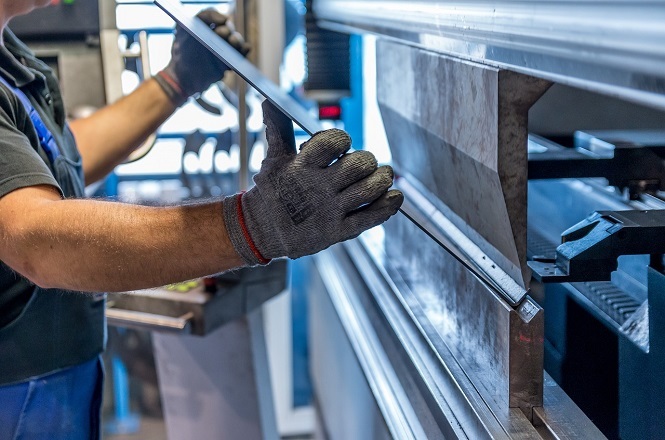 Um trabalhador está operando uma máquina de dobra de metal. Ele está usando luvas de proteção enquanto segura uma chapa de metal que está sendo dobrada pela máquina. A máquina é grande e parece ser feita de metal, com um design industrial. O trabalhador está vestindo uma camisa escura e um avental azul. A cena parece estar em um ambiente de fábrica ou oficina, com iluminação suficiente para o trabalho.