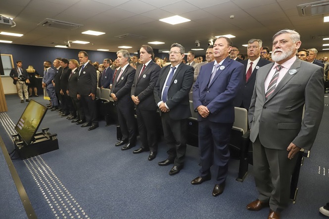 Um grupo de homens de terno está em pé em uma sala de conferências. Eles estão alinhados em fileiras, todos usando trajes formais, como ternos e gravatas. Alguns deles estão usando crachás redondos na lapela. A sala tem um carpete azul e cadeiras cinza. Há um monitor grande no chão à esquerda da imagem. Ao fundo, mais pessoas estão sentadas, e algumas estão em pé.