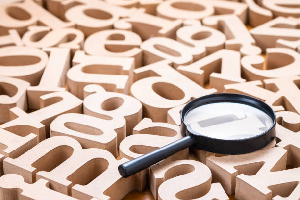 Letras de madeira espalhadas aleatoriamente sobre uma superfície, criando um padrão caótico. No centro da imagem, há uma lupa posicionada sobre algumas das letras, destacando-as. A lupa tem uma alça preta e uma lente circular. As letras são de tamanhos variados e estão em diferentes orientações.