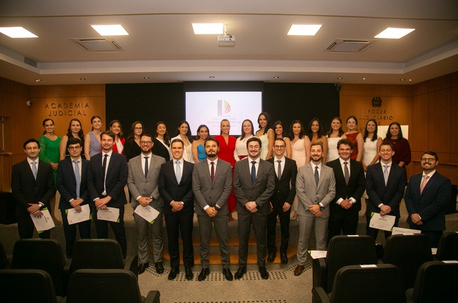 Um grupo de pessoas posando para uma foto em um auditório. Na frente, há alguns homens segurando certificados. Atrás deles, há também um grupo de mulheres, a maioria vestindo roupas formais em tons variados, incluindo vermelho, preto e branco. Ao fundo, há um telão com um logotipo e as palavras "Academia Judicial" e "Poder Judiciário" nas paredes laterais. O ambiente é bem iluminado, com cadeiras de auditório vazias na parte inferior da imagem.