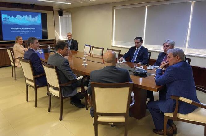 Sala de reunião com sete homens sentados ao redor de uma mesa retangular de madeira. Todos estão vestidos formalmente com ternos. Na parede ao fundo, há uma tela de apresentação que exibe o texto "PROJETO JURISPRUDÊNCIA" e "TRIBUNAL DE CONTAS DO ESTADO". A sala tem janelas com persianas fechadas e o piso é de cor clara. Há copos de água sobre a mesa.