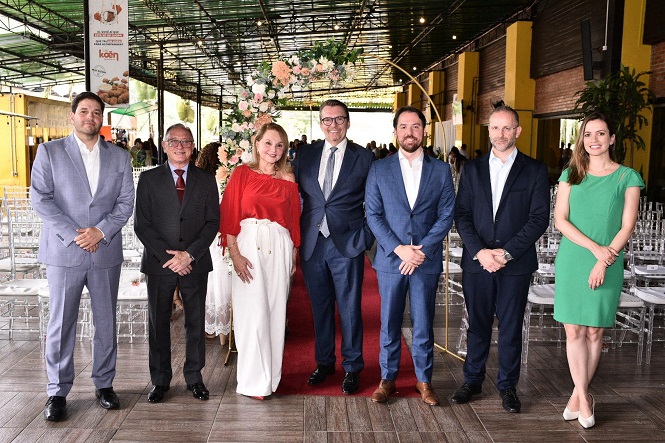 Sete pessoas posando para uma foto em um ambiente que parece ser um salão de eventos. No centro, há um arco decorado com flores, e o chão é coberto por um tapete vermelho. As pessoas estão vestidas formalmente: quatro homens de terno e gravata, uma mulher de blusa vermelha e saia branca, um homem de terno sem gravata e uma mulher de vestido verde. Ao fundo, há cadeiras transparentes organizadas em fileiras, sugerindo que o local está preparado para um evento ou cerimônia. As paredes são amarelas e há plantas decorativas.