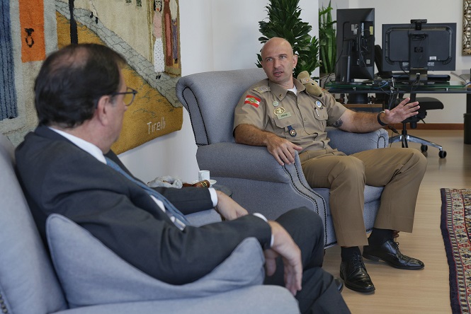 Dois homens estão sentados em poltronas em uma sala. O homem à direita está usando um uniforme militar bege, com várias insígnias e distintivos no peito, e está gesticulando com a mão direita enquanto fala. Ele também tem uma bandeira do Brasil no braço esquerdo. O homem à esquerda está de terno escuro, camisa branca e gravata, e está olhando para o homem de uniforme. Ao fundo, há uma tapeçaria colorida na parede e uma mesa com dois monitores de computador. Há também plantas decorativas na sala e um tapete no chão.