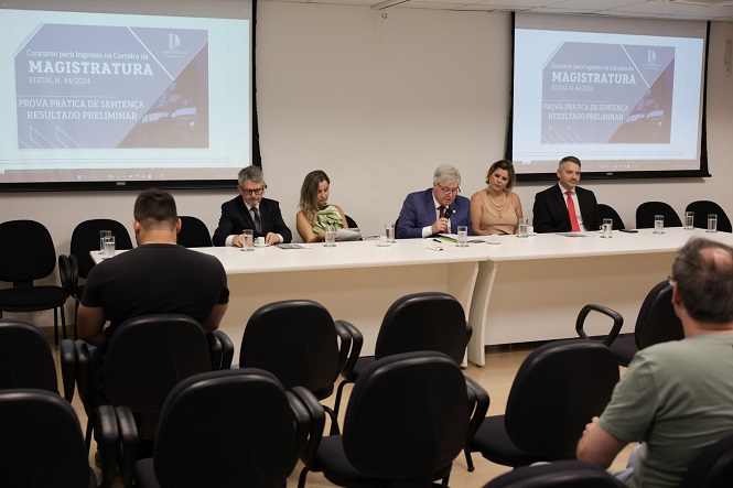 Cinco pessoas estão sentadas lado a lado em uma mesa comprida em uma sala de conferências. À frente delas, há várias cadeiras, algumas ocupadas por pessoas. Na parede ao fundo, há dois projetores exibindo a mesma imagem, que contém o texto: "Concurso para Ingresso na Carreira da Magistratura Edital n. 02/2024 Prova Prática de Sentença Resultado Preliminar". As pessoas na mesa estão vestidas formalmente, e uma delas está falando ao microfone. A sala tem um ambiente profissional e formal.