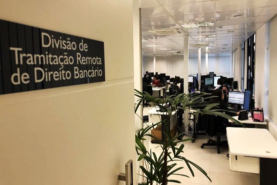 Uma sala de escritório com várias mesas e computadores. Na porta, há uma placa com o texto "Divisão de Tramitação Remota de Direito Bancário". Dentro da sala, várias pessoas estão trabalhando em seus computadores. Há também algumas plantas decorativas no ambiente. A iluminação é clara, com luzes fluorescentes no teto. As janelas têm persianas verticais.