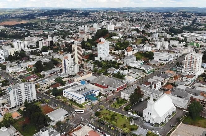 Vista aérea de uma cidade com várias edificações. Há prédios de diferentes alturas, com alguns edifícios altos se destacando. No centro da imagem, há uma igreja branca com uma torre, cercada por áreas verdes. As ruas são bem definidas e há veículos circulando. A cidade está situada em uma área com relevo levemente ondulado, e ao fundo, é possível ver colinas e vegetação. O céu está parcialmente nublado.