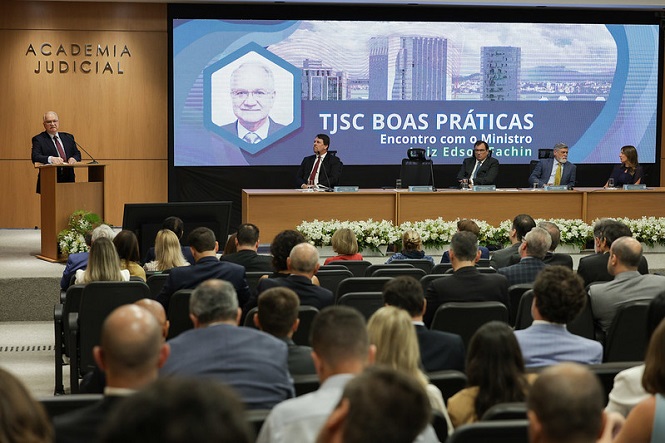 Auditório com várias pessoas sentadas assistindo a uma apresentação. À esquerda, um homem de terno está em um púlpito falando ao microfone. No centro, há uma grande tela de projeção com a imagem de um homem idoso e a seguinte inscrição: "TJSC BOAS PRÁTICAS Encontro com o Ministro Luiz Edson Fachin". Ao fundo, quatro pessoas estão sentadas em uma mesa de conferência, olhando para a plateia. O ambiente é formal, com todos vestidos de maneira elegante, e há arranjos de flores brancas decorando a mesa. Na parede à esquerda, está escrito "ACADEMIA JUDICIAL".