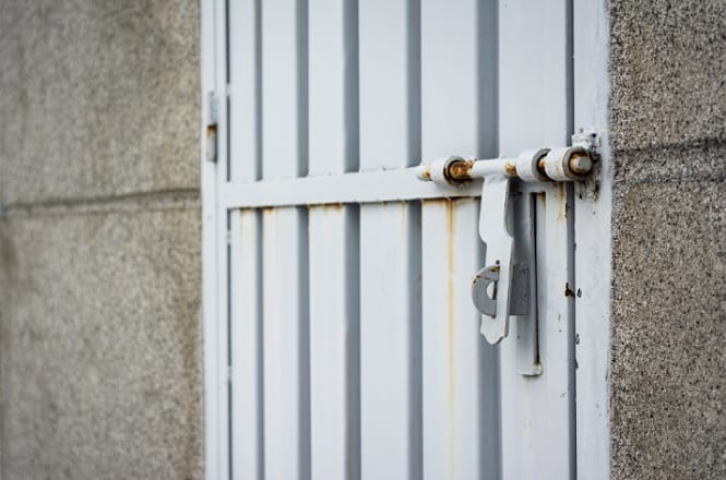 Portão de metal cinza com um cadeado pendurado. O portão tem barras verticais e está fixado a uma parede de concreto. Há sinais de ferrugem nas dobradiças e na área ao redor do cadeado. A parede de concreto tem uma textura áspera e está dividida por uma linha horizontal.