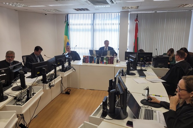 Sala de tribunal com várias pessoas sentadas em torno de mesas brancas, cada uma equipada com computadores e monitores. Há seis pessoas visíveis, todas vestidas formalmente, algumas usando togas. No centro da sala, há uma mesa com vários livros empilhados e duas bandeiras ao fundo, uma do Brasil e outra vermelha. As janelas ao fundo têm persianas verticais que permitem a entrada de luz natural. O ambiente parece ser de um julgamento ou audiência judicial.