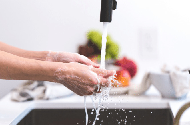 Uma pessoa está lavando as mãos em uma pia. A água está fluindo de uma torneira preta, e há bastante sabão nas mãos da pessoa, formando espuma. Ao fundo, desfocado, é possível ver algumas frutas em uma tigela, incluindo algo que parece ser maçãs e talvez uma alface. A cena transmite uma sensação de limpeza e higiene.